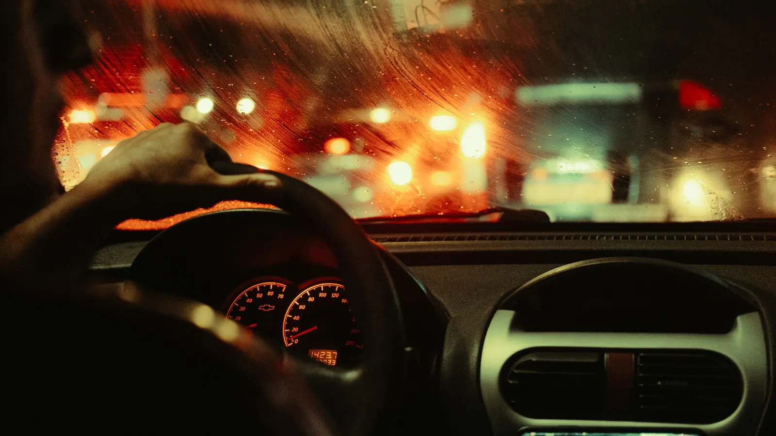 Manos sobre el volante de un auto en marcha de noche, con las luces de la ciudad a través del parabrisas
